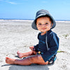 A cute baby boy sitting on a white sandy beach with the navy pinstripe Bucket Sun Protection Hat squinting at the viewer.