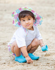 A cute young girl wearing Brim Sun Protection Hat while squatting and playing in the sand. She is looking at the viewer with a questionable smirk.