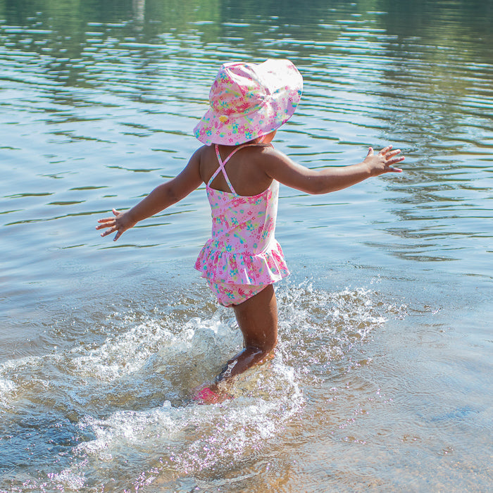 A cute young girl skipping away into the water towards the camera while wearing the light pink dragonfly floral Brim Sun Protection Hat and a matching swimsuit.