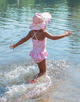 A cute young girl skipping away into the water towards the camera while wearing the light pink dragonfly floral Brim Sun Protection Hat and a matching swimsuit.