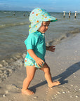 A young toddler walking along the shore with her light Aqua Sea Friends Flap Sun Protection Hat and a swimming outfit to match.