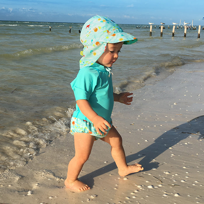 A young toddler walking along the shore with her light Aqua Sea Friends Flap Sun Protection Hat and a swimming outfit to match.