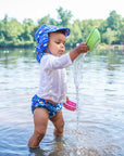 A young boy watching the water pour out of a toy boat he is holding up while standing in a lake and wearing the Royal Blue Sea Friends Flap Sun Protection Hat.