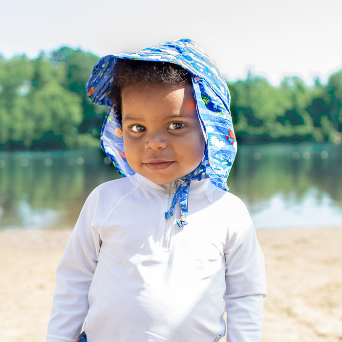 A young boy looking intently at the viewer with a little smile and his Royal Blue Sea Friends Flap Sun Protection Hat on.