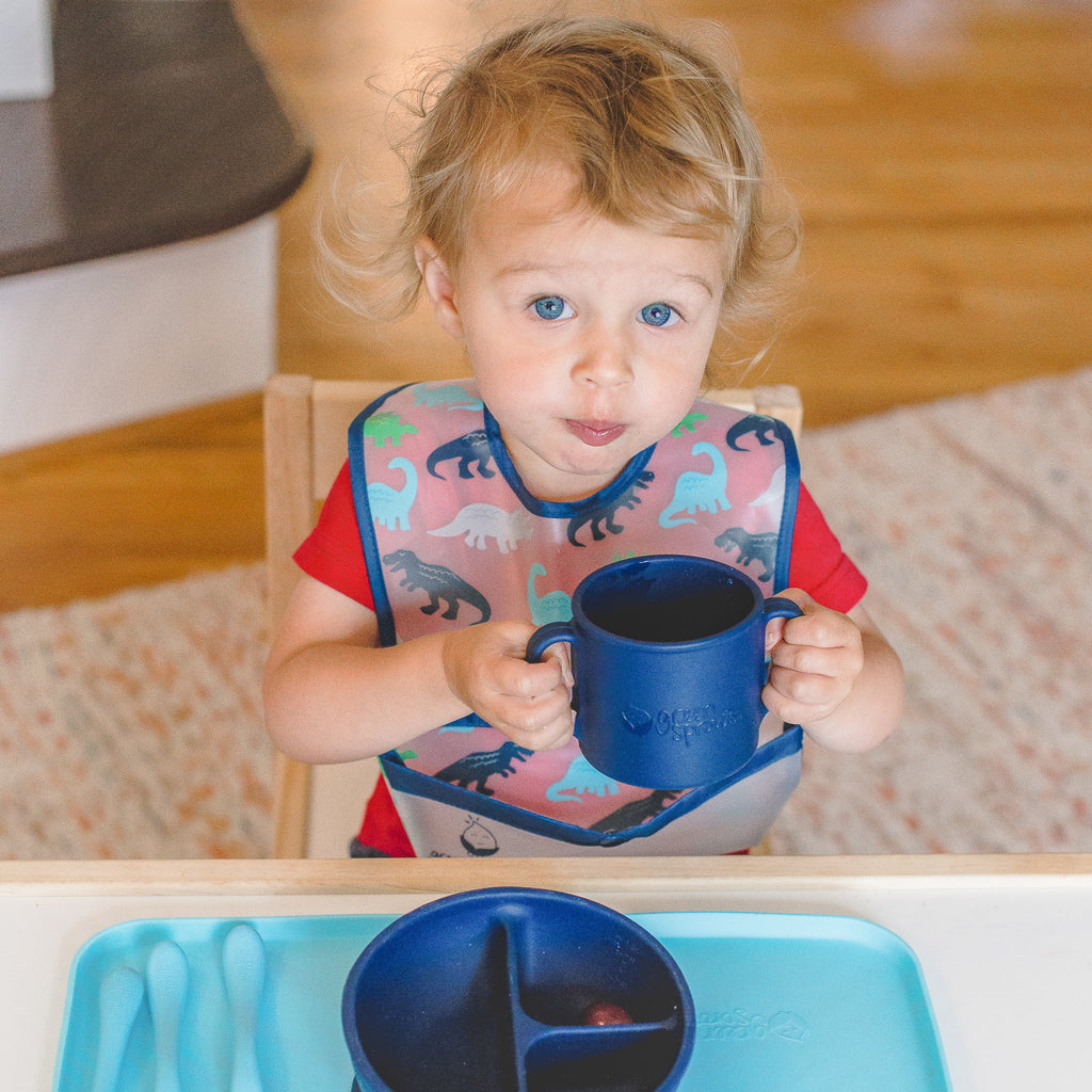 A young toddler holding in a big gulp in his cheeks while holding the navy Learning Cup made from Silicone
