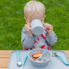 A little boy sitting at a table outside while drinking out of the gray Learning Cup made from Silicone
