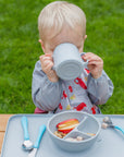 A little boy sitting at a table outside while drinking out of the gray Learning Cup made from Silicone