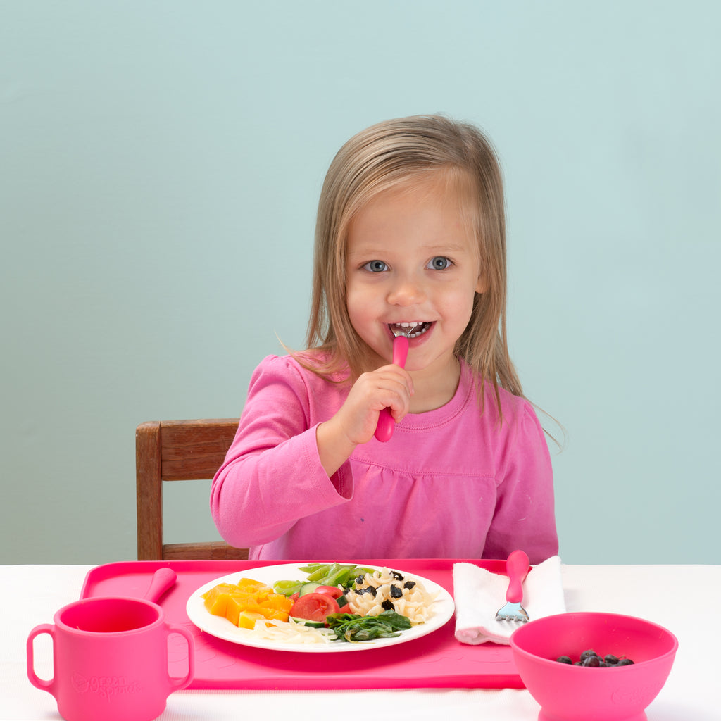 A young girl eating with matching pink dishes and utensils along with a pink Feeding Bowl made from Silicone with raisins inside.