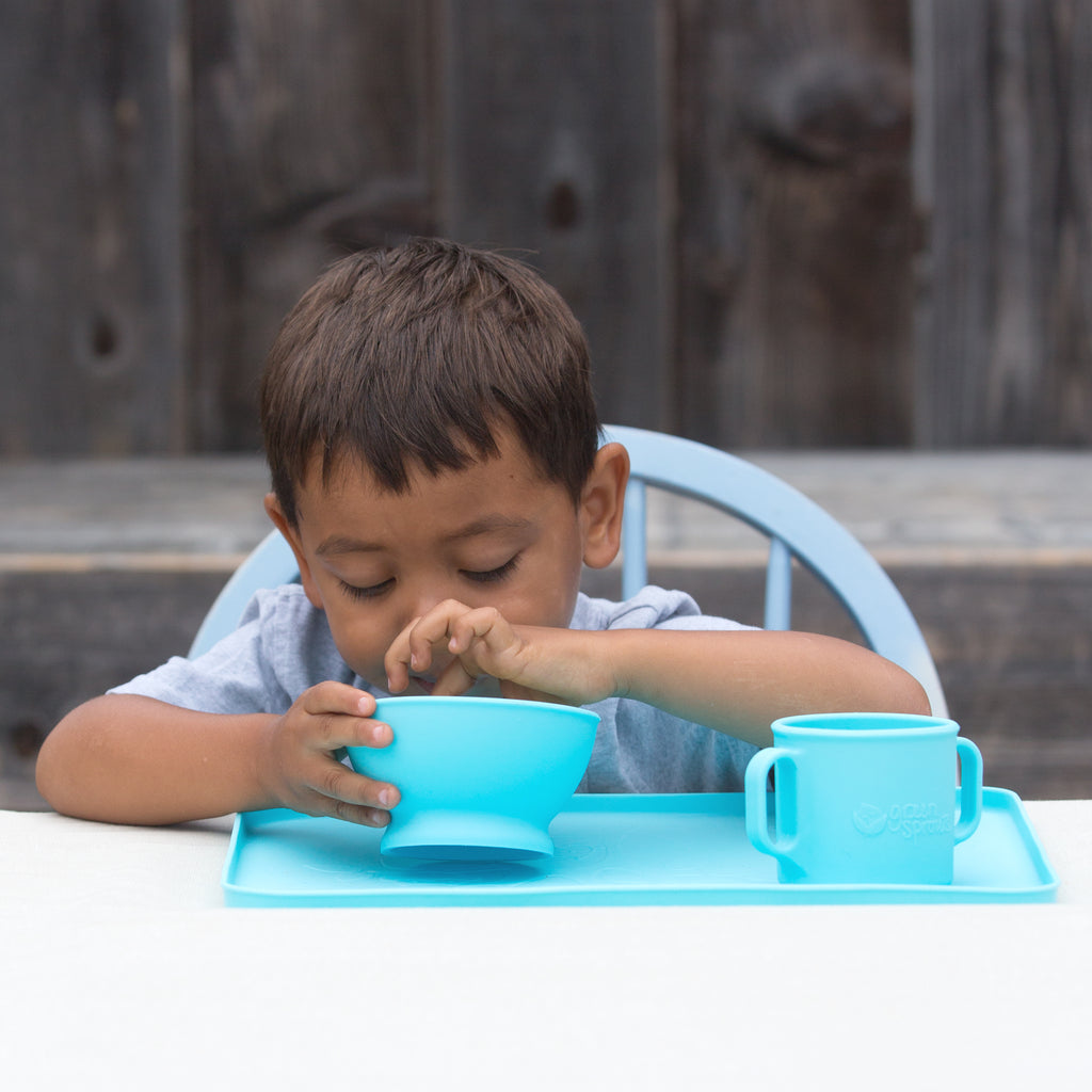 A young boy eating out of an Aqua Feeding Bowl made from Silicone with a matching aqua cup and platemat in a backyard with a wooden fence behind him.