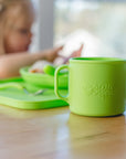 A close up of the green Learning Cup made from Silicone with a little girl eating in the background