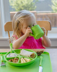 A little girl drinking out of the green Learning Cup made from Silicone and sitting with the rest of her green dinner wear