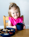 A cute little girl smirking while holding her navy Learning Cup made from Silicone