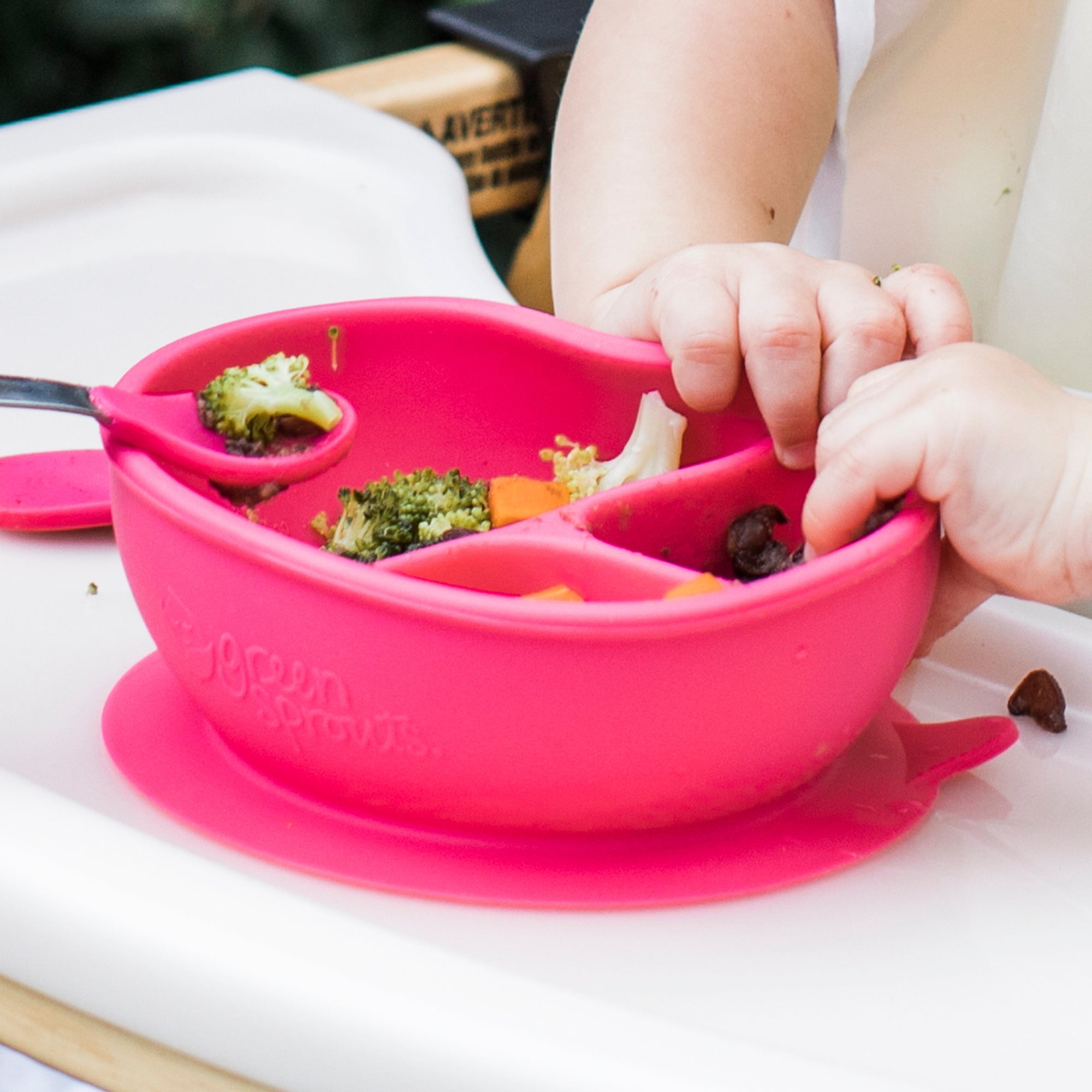 A close up of the pink Learning Bowl made from Silicone with food in it and a baby&#39;s hands bending the bowl.