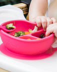 A close up of the pink Learning Bowl made from Silicone with food in it and a baby's hands bending the bowl.
