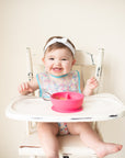 A giggling toddler girl sitting in her high chair with the pink Learning Bowl made from Silicone