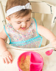 A little girl sitting in a white wooden high chair using a learning spoon to pick up her food from the pink Learning Bowl made from Silicone