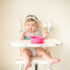 A giggling toddler girl sitting in her high chair with the Pink Learning Spoon Set