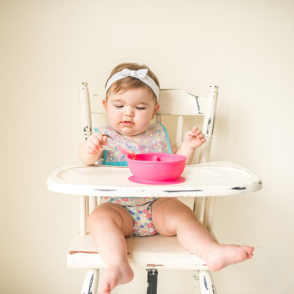 A giggling toddler girl sitting in her high chair with the Pink Learning Spoon Set