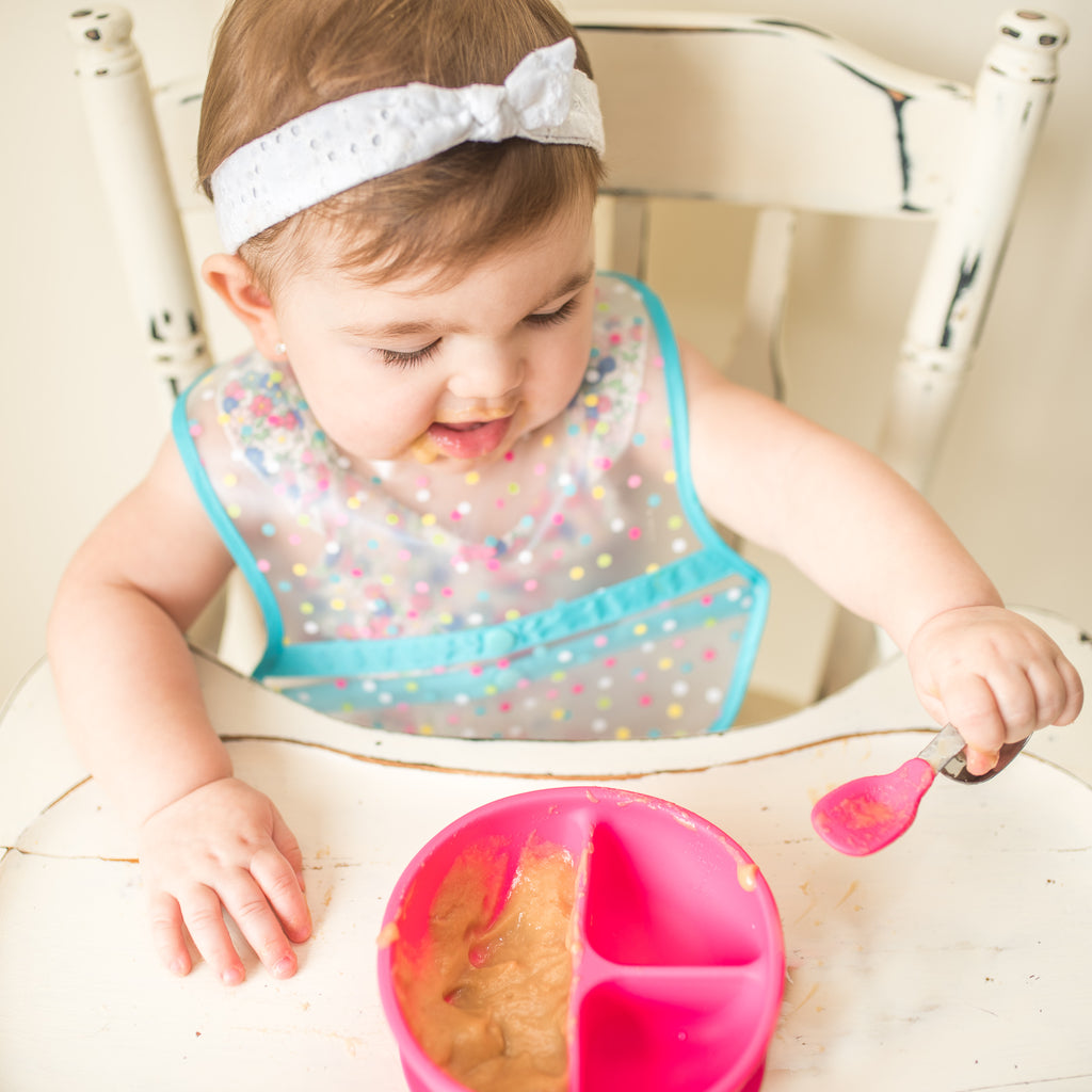 A little girl sitting in a white wooden high chair picking up her food with the Pink Learning Spoon Set