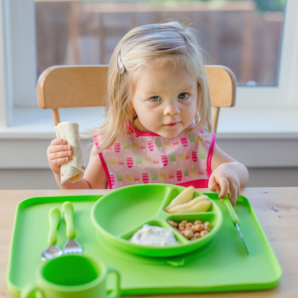 A young girl holding a burrito in one hand and the green Learning Cutlery Set in the other