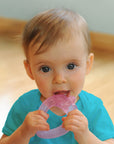 A wide-eyed 6 month old girl looking intently while gripping the light pink strawberry Cool Fruit Teether to her little mouth.