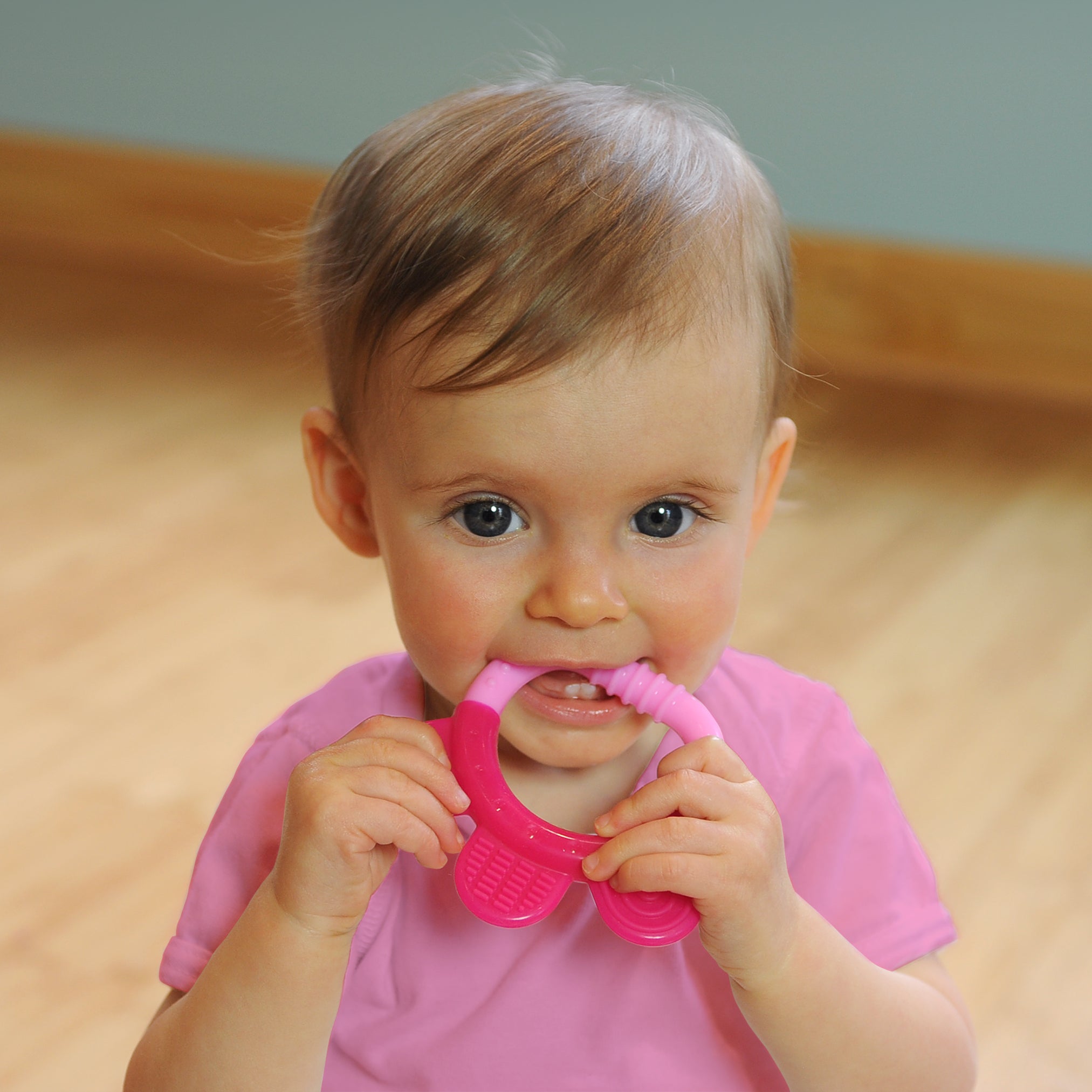 A wide-eyed 6 month old girl looking intently while gripping the pink Everyday Teether made from Silicone to her little mouth.