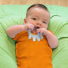 A little newborn boy laying on a green blanket and playing with the light blue cooling everyday teether in his mouth.