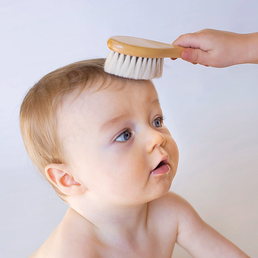 A toddler looking up at someone who is brushing his hair with the baby brush.