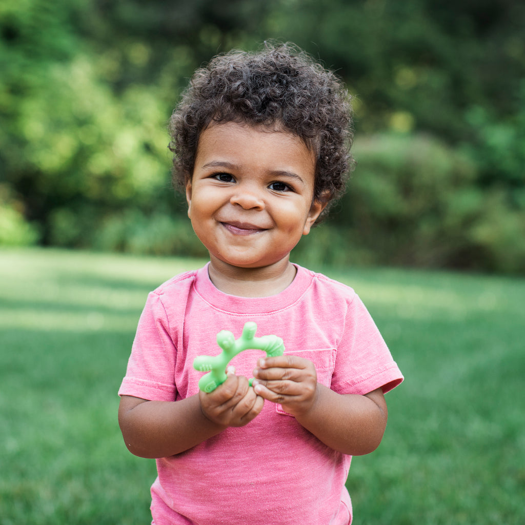 Toddler in a red shirt standing outside grinning at the viewer and holding a Front and Side Teether made from Silicone.
