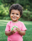 Toddler in a red shirt standing outside grinning at the viewer and holding a Front and Side Teether made from Silicone.