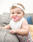A cute infant girl playfully smiling and chewing one a pink first teether made from silicone while standing on a couch and pillows.