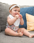A little girl sitting on a gray couch and looking to the side while holding a pink first teether made from silicone up to her mouth.