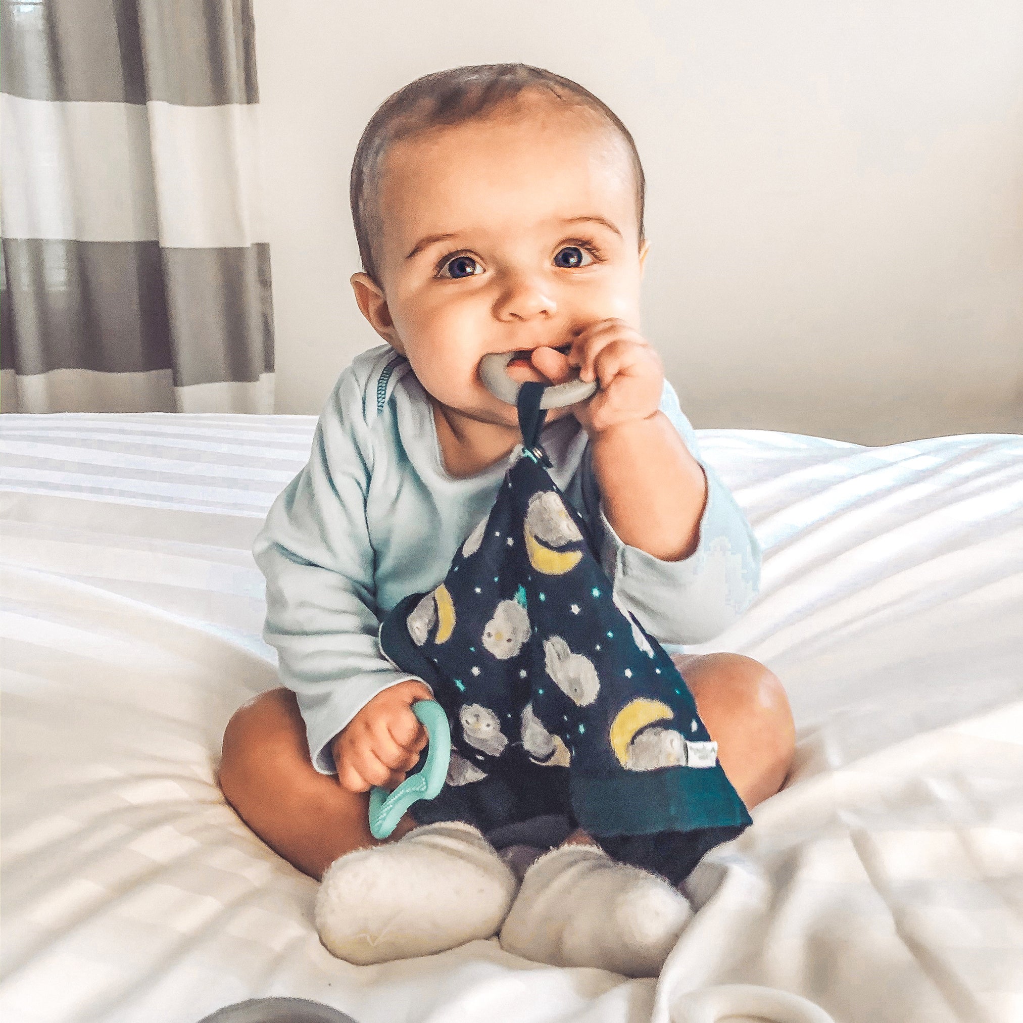 An adorable infant boy with long eyelashes looking up is hold an aqua teether in one hand a gray first teether made from silicone in his other hand up to his mouth.