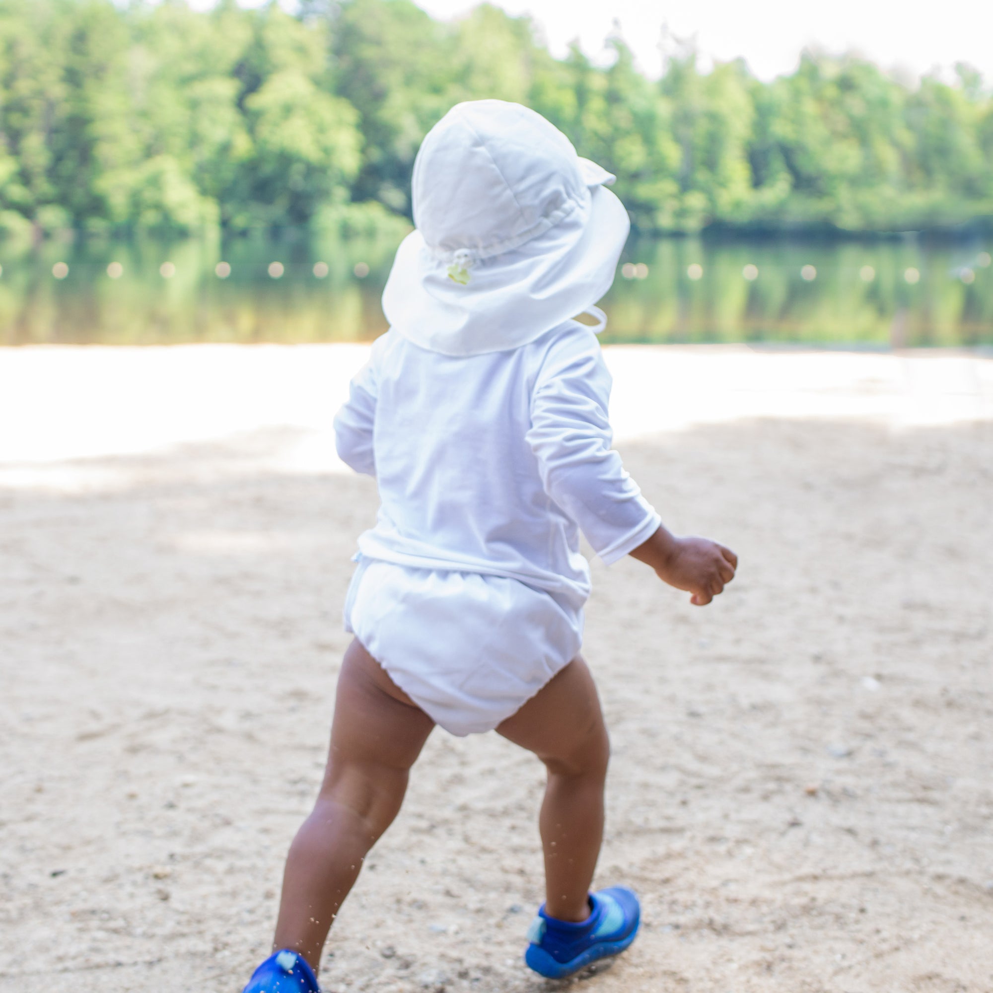 A little boy waddling away on the beach while wearing a white swim outfit including a white Flap Sun Protection Hat.