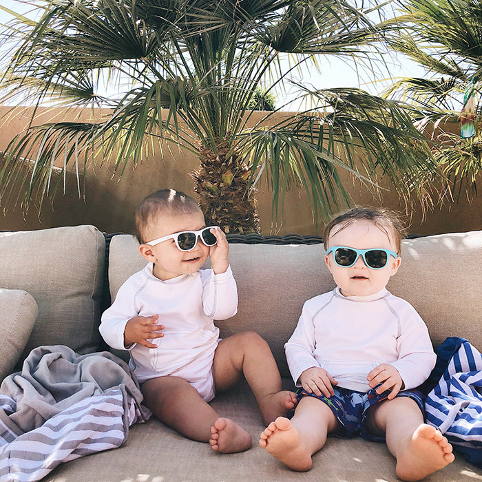 Two babies smiling while on a couch under a palm tree with the white Breathable Sun Protection Shirt on.