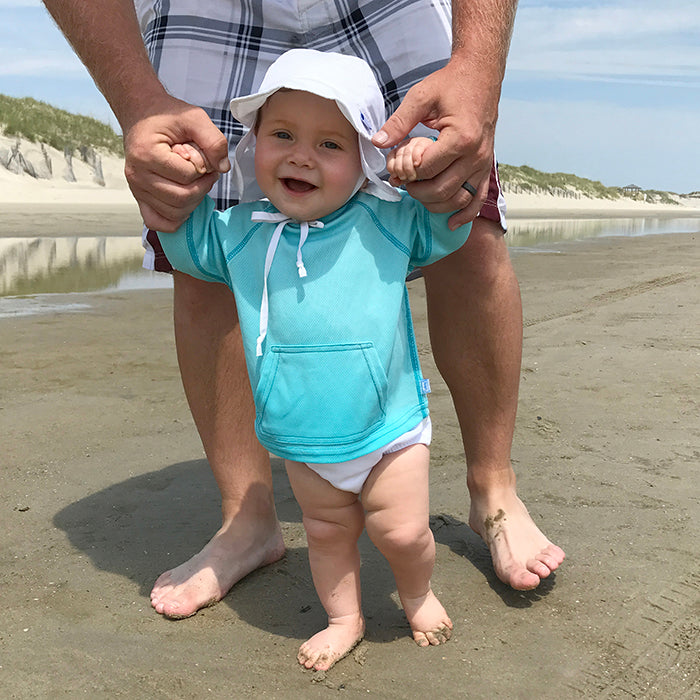 A smiling baby wearing the aqua Breathable Sun Protection Shirt while learning to walk and holding her parent&#39;s hand on the beach.