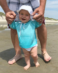 A smiling baby wearing the aqua Breathable Sun Protection Shirt while learning to walk and holding her parent's hand on the beach.