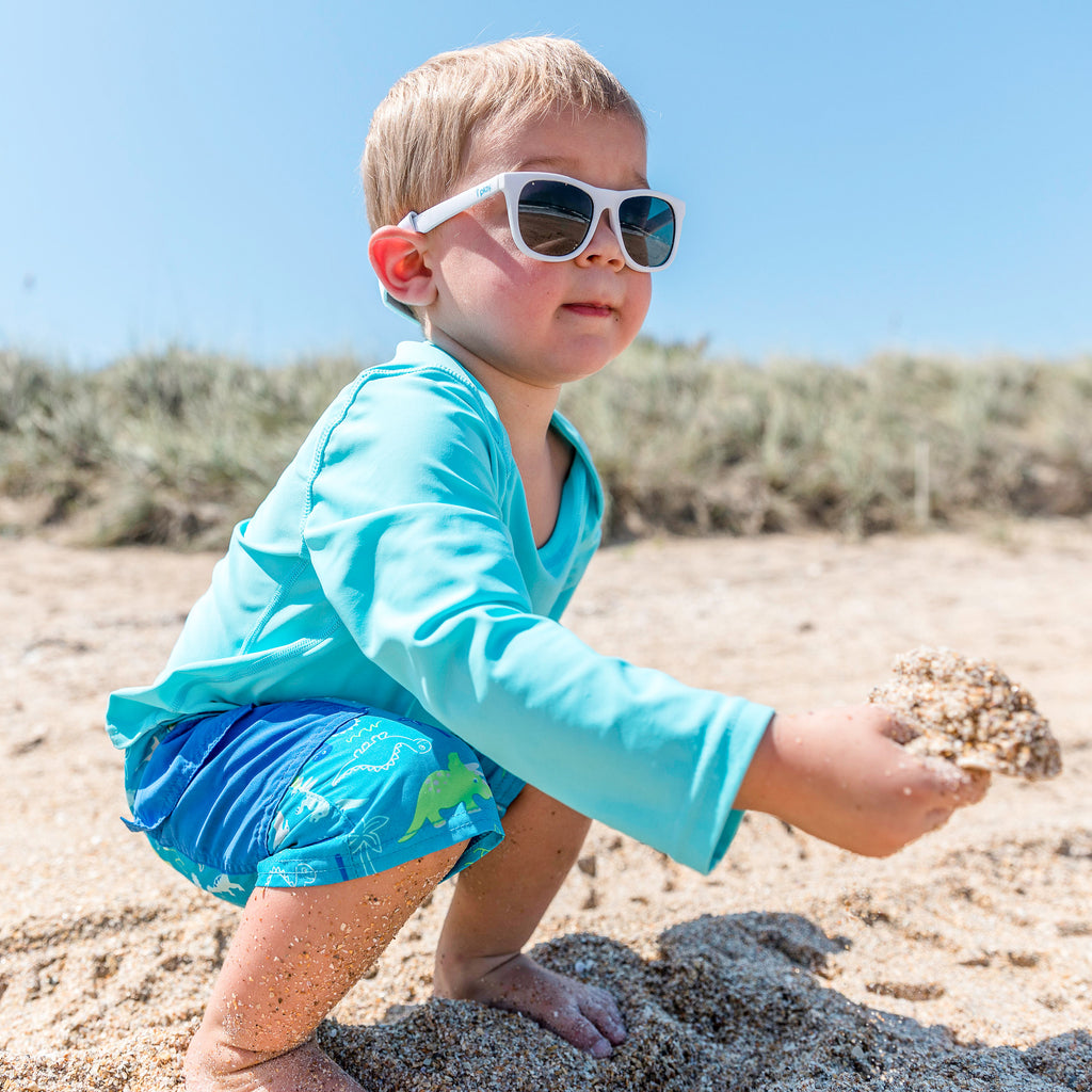 A cute little dude squatting on the beach and picking up sand with a seashell like a tortia chip and queso. He rocking some white Flexible Sunglasses and aqua swim outfit.