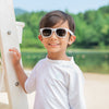 An young boy with an adorably forced smile posing with his arm on a lifeguard pole with some white Flexible Sunglasses on.