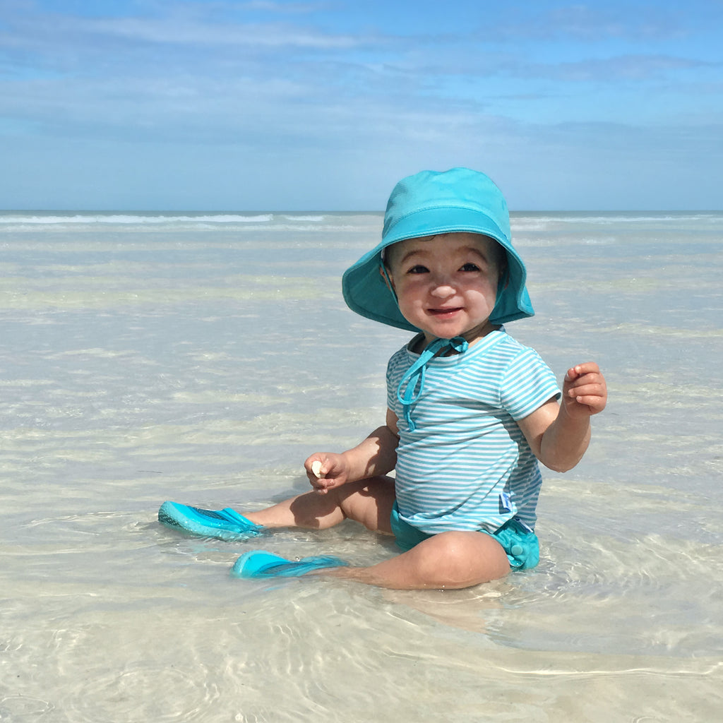 A little girl sitting in the shallow water on the beach while wearing a light aqua pinstripe Cap Sleeve Rashguard Shirt